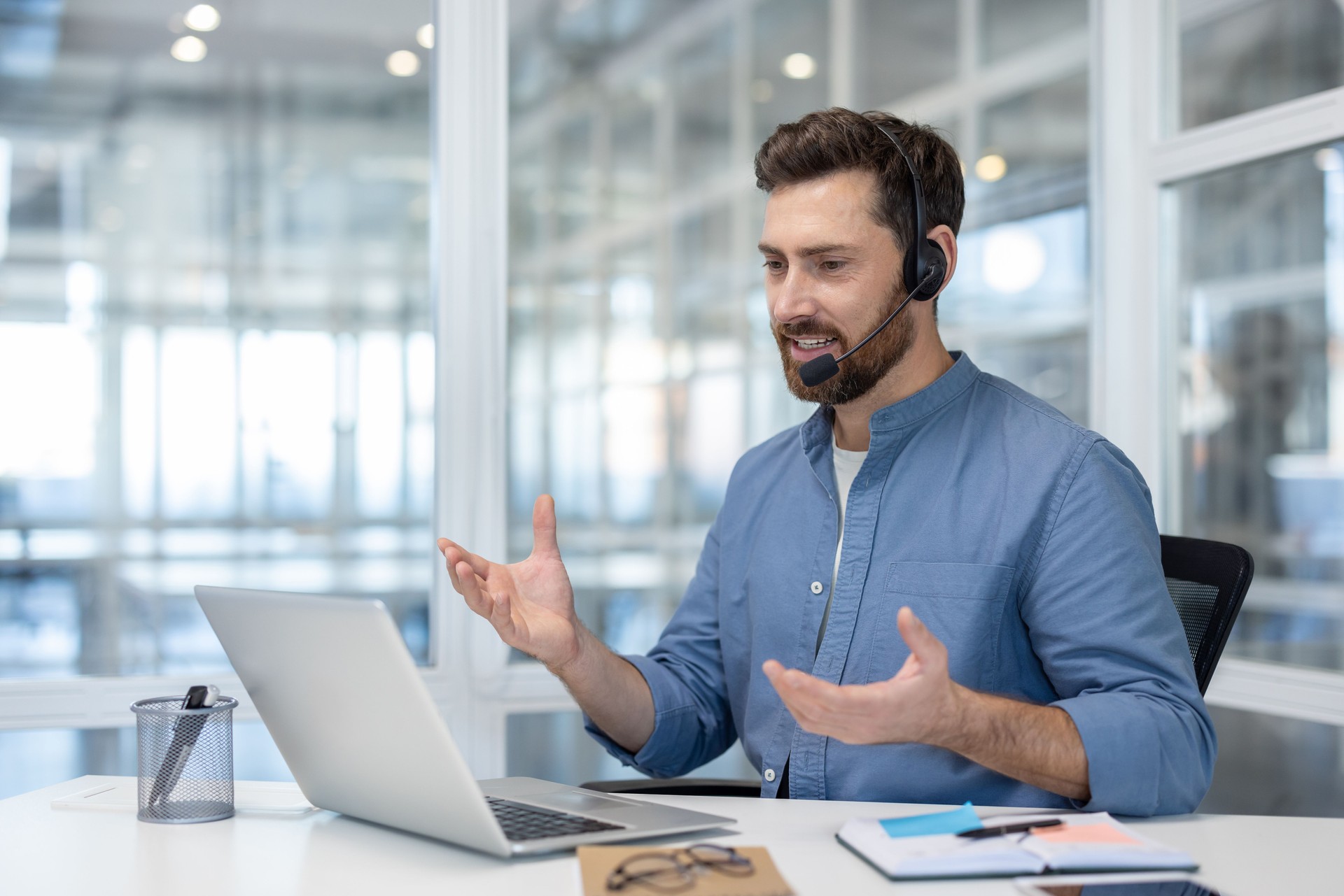 Young man with headset talking to colleagues and partners remotely using laptop for video call. Support worker, helpline consultant inside office